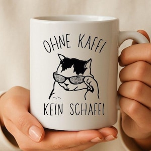 Könnte beinhalten: Weißer Keramikbecher mit einer schwarz-weißen Illustration einer Katze mit Sonnenbrille. Der Becher trägt den Text "OHNE KAFFI" über der Katze und "KEIN SCHAFFI" darunter, in schwarzer Schrift. Der Becher wird von einer Person gehalten.