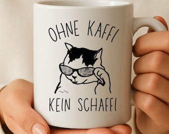 May include: White ceramic mug featuring a black and white illustration of a cat wearing sunglasses. The mug has the text "OHNE KAFFI" above the cat and "KEIN SCHAFFI" below it, in black lettering. The mug is held by a person.