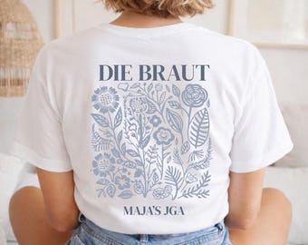 May include: White t-shirt with the text "DIE BRAUT" at the top and "MAJA'S JGA" at the bottom. The shirt features a floral design in shades of blue. The back of the shirt is visible.