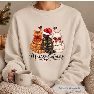 May include: A beige crewneck sweatshirt featuring a graphic of three cats sitting side-by-side, adorned with red bows and a string of Christmas lights. The center cat wears a red Santa hat. Below the cats, the text reads Merry Catmas.
