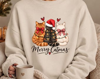 May include: A beige crewneck sweatshirt featuring a graphic of three cats sitting side-by-side, adorned with red bows and a string of Christmas lights. The center cat wears a red Santa hat. Below the cats, the text reads Merry Catmas.