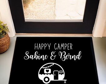May include: Black rectangular doormat with white text that reads "HAPPY CAMPER Sabine & Bernd". A white graphic of a camper is below the text. The doormat is placed in front of a black door.