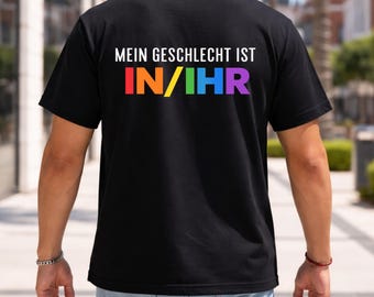 May include: A black short-sleeved t-shirt featuring the white text MEIN GESCHLECHT IST on the back. Below this, the text IN/IHR is printed in a rainbow gradient of red, orange, yellow, green, blue, and purple block letters on the fabric.