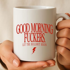 Könnte beinhalten: Ein weißer Keramikbecher mit rotem Text, der GOOD MORNING FUCKERS. LET THE BULLSHIT BEGIN. lautet, sowie einem kleinen roten Blitzsymbol unter dem Text. Der Becher hat einen Standardhenkel und ein glattes, zylindrisches Design.
