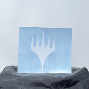 May include: A light blue, rectangular block with a white, stylized crown emblem. The block is resting on a dark, textured surface against a white background. The emblem is a prominent design element.