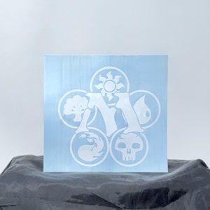 May include: A light blue square block with a white Magic: The Gathering symbol. The symbol features a stylized letter "M" surrounded by five circular icons representing different elements. The block is resting on a dark gray surface.