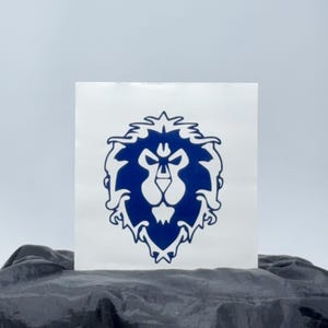 May include: A blue and white sticker featuring the Alliance emblem from the video game World of Warcraft. The emblem is a stylized lion's head with a detailed mane and facial features. The sticker is displayed on a white background.