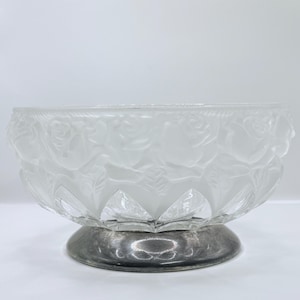 May include: A clear glass bowl with a frosted rose design around the rim, set on a silver-colored pedestal base. The bowl's sides are faceted, and the base is round. The overall design is elegant and decorative.