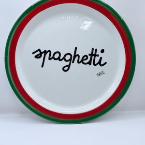May include: White ceramic spaghetti plate with a green and red rim. The word "spaghetti" is written in black cursive in the center of the plate. A signature is visible near the bottom right.