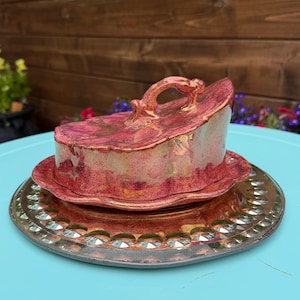 May include: A decorative, antique-style butter dish with a lid and handle. The dish is a deep red with gold accents, sitting on a matching plate with a decorative edge. The set is placed on a light blue surface.