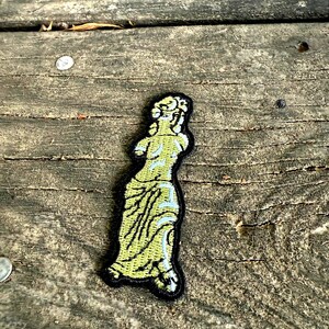 May include: Embroidered patch featuring a classical statue design. The patch depicts a light green figure with blue accents and a black border. The patch is placed on a weathered wooden surface.