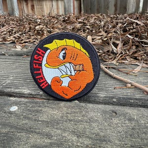 May include: A round, black patch with the text "HELLFISH" in red. The patch features an orange cartoon fish with a cigar, flexing its arm. The fish has a yellow fin and white teeth.
