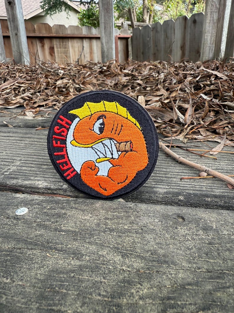 May include: A round, black patch with the text "HELLFISH" in red. The patch features an orange cartoon fish with a cigar, flexing its arm. The fish has a yellow fin and white teeth.