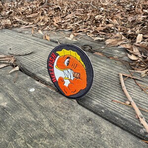 May include: Embroidered patch featuring a cartoonish orange fish with yellow fins and sharp teeth, holding a cigarette. The patch has the word "HELLFISH" in red letters. The patch is on a black background with a white border.