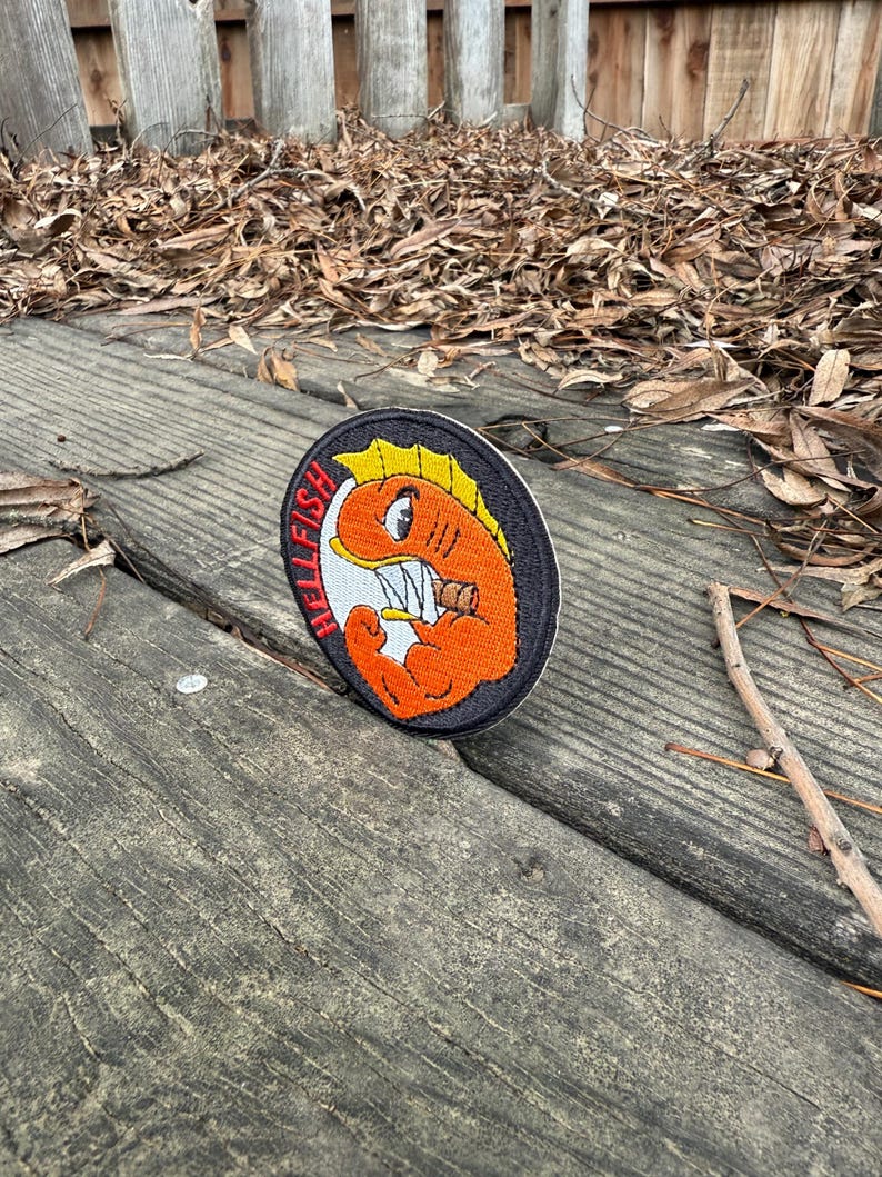 May include: Embroidered patch featuring a cartoonish orange fish with yellow fins and sharp teeth, holding a cigarette. The patch has the word "HELLFISH" in red letters. The patch is on a black background with a white border.