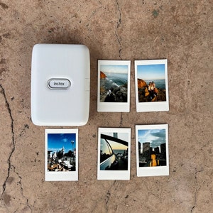 Puede incluir: Impresora de c&aacute;mara instant&aacute;nea blanca con la palabra "instax" impresa en la parte delantera. La impresora est&aacute; rodeada de seis fotos instant&aacute;neas con varias im&aacute;genes.
