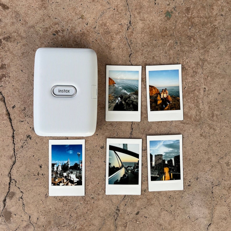 Puede incluir: Impresora de c&aacute;mara instant&aacute;nea blanca con la palabra "instax" impresa en la parte delantera. La impresora est&aacute; rodeada de seis fotos instant&aacute;neas con varias im&aacute;genes.