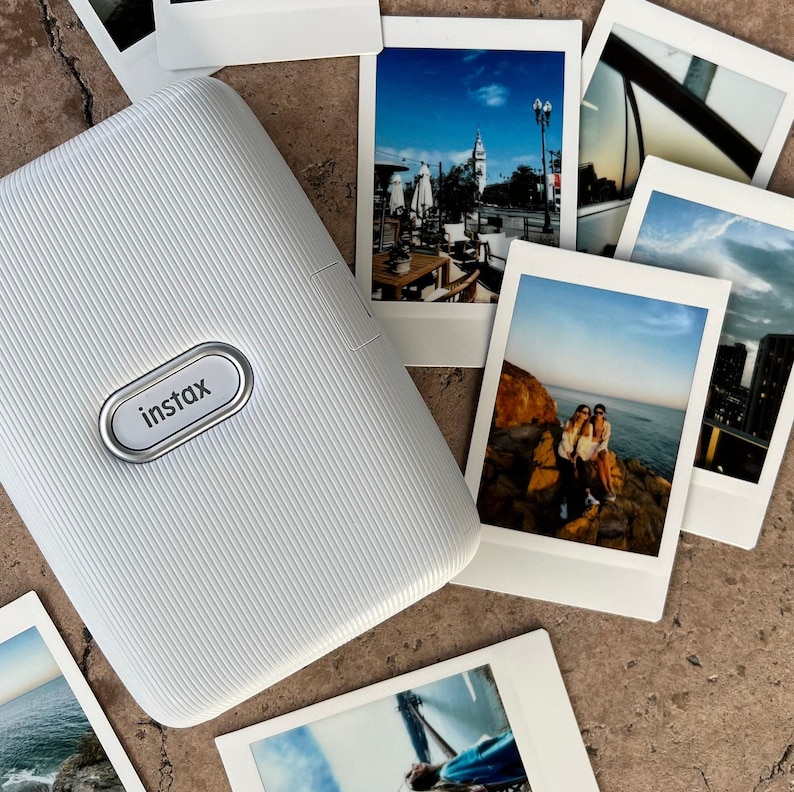 Puede incluir: Impresora de c&aacute;mara instant&aacute;nea blanca con la palabra "instax" impresa en la parte delantera. La impresora est&aacute; rodeada de varias fotos instant&aacute;neas.
