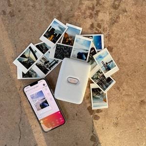 Puede incluir: Una impresora de fotos port&aacute;til blanca con una pantalla que muestra una foto de una playa. La impresora est&aacute; rodeada de fotos instant&aacute;neas de varios paisajes y paisajes urbanos.