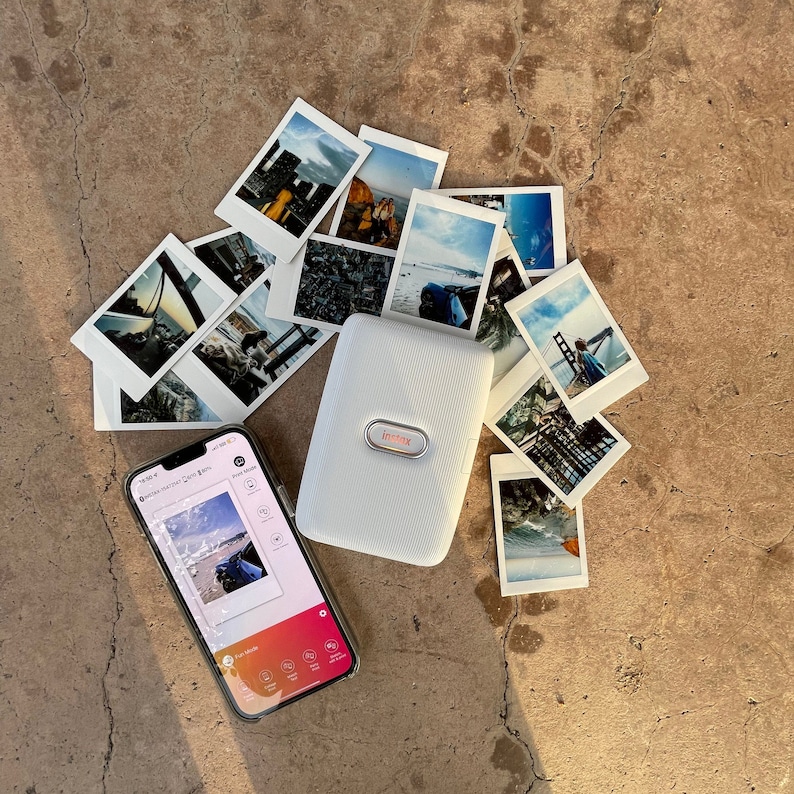 Puede incluir: Una impresora de fotos port&aacute;til blanca con una pantalla que muestra una foto de una playa. La impresora est&aacute; rodeada de fotos instant&aacute;neas de varios paisajes y paisajes urbanos.
