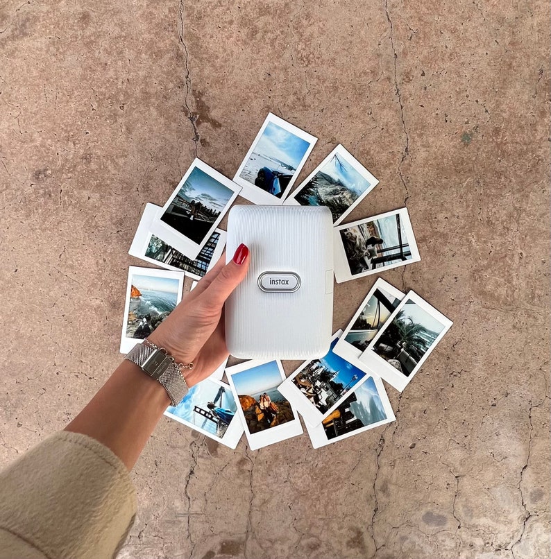 Puede incluir: Una impresora de c&aacute;mara instant&aacute;nea blanca con la palabra "instax" impresa en ella. La c&aacute;mara est&aacute; rodeada de fotos instant&aacute;neas con varios paisajes y paisajes urbanos.