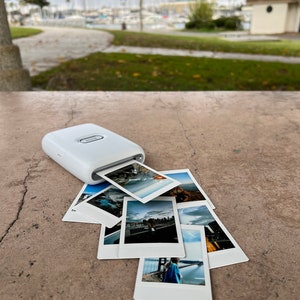 Puede incluir: Una impresora de fotos port&aacute;til blanca con una pila de fotos impresas sobre una superficie de hormig&oacute;n. Las fotos son de varios paisajes y paisajes urbanos.