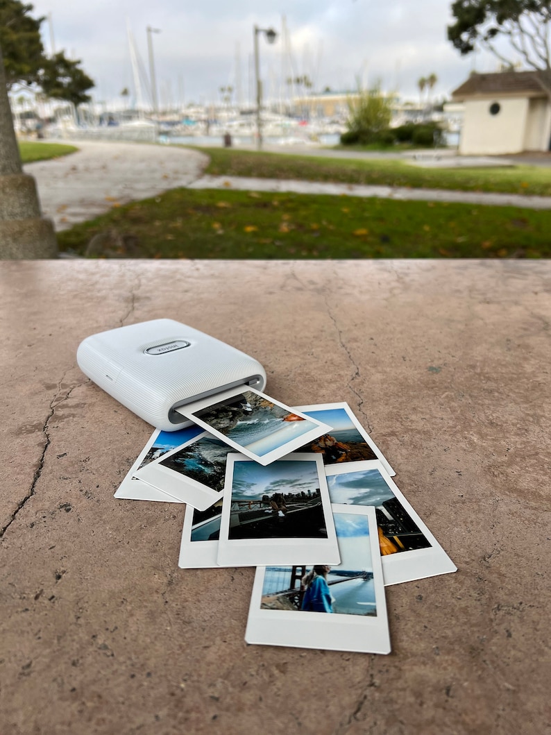 Puede incluir: Una impresora de fotos port&aacute;til blanca con una pila de fotos impresas sobre una superficie de hormig&oacute;n. Las fotos son de varios paisajes y paisajes urbanos.