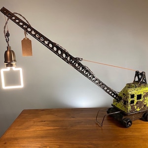 May include: A vintage toy crane lamp with a yellow and green paint job. The crane is made of metal and has a working light bulb. The lamp is on a wooden table.