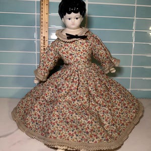May include: A vintage porcelain doll with a black bob hairstyle and a floral print dress. The dress features long sleeves, a lace collar, and a black bow. The doll is wearing black shoes and has a cream-colored lace trim on the dress.