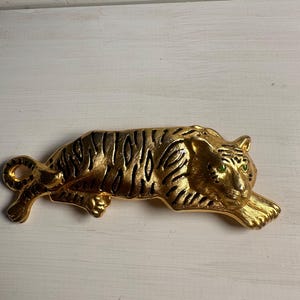 May include: A gold-toned metal brooch shaped like a reclining tiger. The tiger features a textured finish with black enamel stripes across its body and two small, bright green stones set as eyes. The animal is depicted in a resting pose.