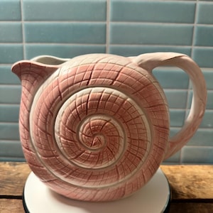 May include: A ceramic pitcher shaped like a spiral seashell with a pink and white gradient finish. The surface features a textured, grid-like pattern that follows the spiral design, a curved handle on the right side, and a small pouring spout.