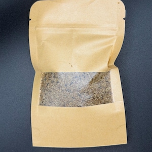May include: A tan paper bag with a clear window, partially open, revealing a granular, brown substance. The bag has a zip closure at the top and is set against a dark background. The contents are visible through the transparent section.