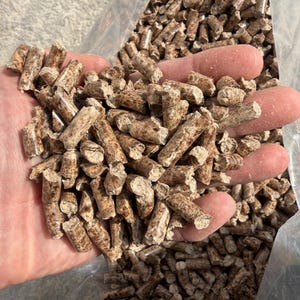 May include: Close-up of wood pellets, cylindrical in shape, with a light brown color and visible wood grain. The pellets are held in a hand, with a clear plastic bag in the background. The pellets are used for grilling and smoking.