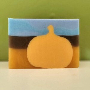 May include: A rectangular bar of soap featuring a layered design with a light blue top section, a dark brown middle band, and a yellow bottom section. A large, solid yellow pumpkin shape is centered on the front of the soap bar.