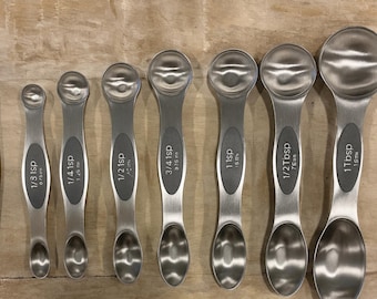 May include: A set of stainless steel measuring spoons and a leveling tool. The spoons are labeled with measurements like 1/3 tsp, 1/4 tsp, 1/2 tsp, 3/4 tsp, 1 tsp, 1/2 Tbsp, and 1 Tbsp. The leveling tool is labeled "LEVELER".