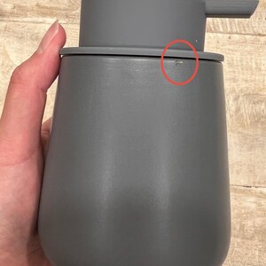 May include: A gray soap dispenser with a pump. The dispenser has a rounded body and a matching gray pump head. The design is minimalist and modern, suitable for a bathroom or kitchen. The dispenser is made of a solid material.