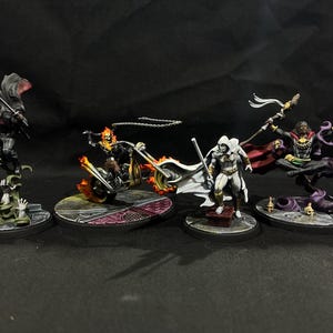 May include: Four miniature figurines of comic book characters. The figures include a black-clad character with a cape, a flaming skull riding a motorcycle, a white-suited character, and a levitating figure with a staff. Each figure is on a circular base.