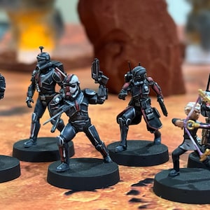 May include: Five black and red painted miniature figures with different weapons and poses, standing on black circular bases. The figures are set against a backdrop of a red rock formation.