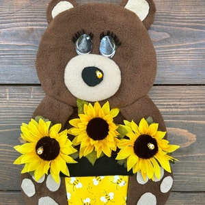 May include: A brown teddy bear holding a yellow flower pot with a bee pattern. The pot has three yellow sunflowers with brown centers and a small yellow bee on each flower.