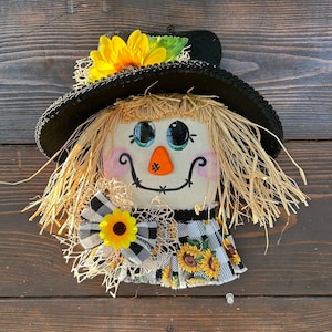 May include: A handmade scarecrow head with a black hat, a yellow sunflower, and a black and white checkered bow. The scarecrow has a painted face with big eyes, a nose, and a stitched smile.