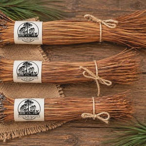 May include: Three bundles of dried brown pine needles, each tied with twine and wrapped in a white paper band featuring a circular logo with the text TOATS MAGOATS. The bundles are arranged horizontally, showcasing the natural texture of the needles.
