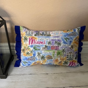 May include: A blue and white pillow with a map of Miami, Florida. The pillow has a blue ruffle around the edges and features the text "Miami" in pink letters.