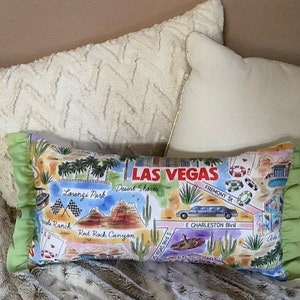 May include: A green and white pillow with a map of Las Vegas, Nevada. The pillow features landmarks like the Fremont Street Experience, the Las Vegas Strip, and the Red Rock Canyon National Conservation Area. The pillow is decorated with a green ruffle.