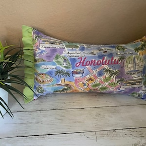 May include: A decorative pillow with a colorful map of Honolulu, Hawaii. The pillow features landmarks like Waikiki Beach, Diamond Head, and Hanauma Bay. The pillow is made of a soft, comfortable fabric and has a green ruffle trim.