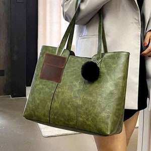 May include: A large, olive green tote bag with long handles and a black pom-pom charm. A small, brown leather wallet is attached to the handle. The bag has a textured appearance and a rectangular shape, suitable for everyday use.
