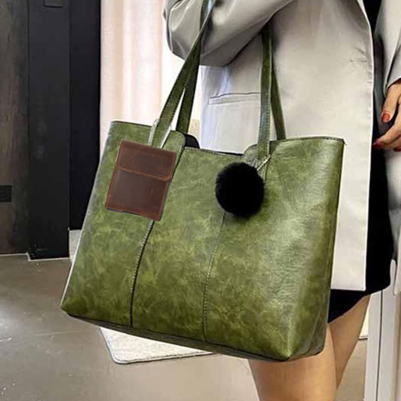 May include: A large, olive green tote bag with long handles and a black pom-pom charm. A small, brown leather wallet is attached to the handle. The bag has a textured appearance and a rectangular shape, suitable for everyday use.