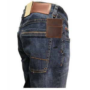 May include: Dark blue denim jeans with a brown leather patch and a small brown leather wallet attached to the belt loop with a silver carabiner. The jeans have orange stitching.