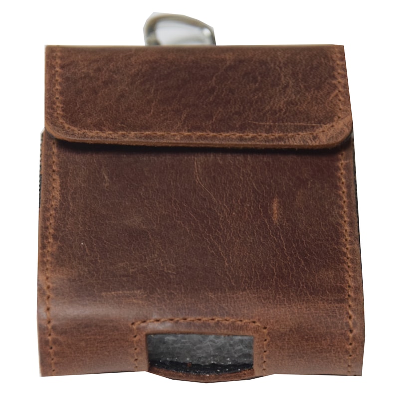 May include: A brown leather case with a flap closure. The case has a rectangular shape with a textured surface and visible stitching. A silver-colored clip is attached to the top. The case is designed to hold a small item.
