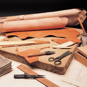 May include: A leather crafting workspace with various tools and materials. Rolled and flat pieces of tan leather are arranged on a work surface. Tools include scissors, a knife, a wooden box, and a compass. A paint can and a book are also present.