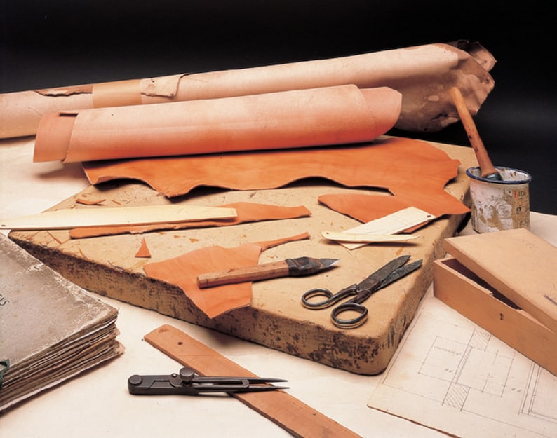 May include: A leather crafting workspace with various tools and materials. Rolled and flat pieces of tan leather are arranged on a work surface. Tools include scissors, a knife, a wooden box, and a compass. A paint can and a book are also present.